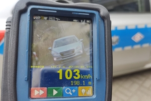 zdjęcie miernika prędkości na którym widać srebrny samochód oraz cyfrę 103